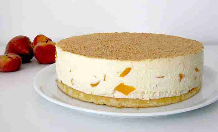 tarta-de-crema-de-queso-batido-con-trocitos-de-melocotón-y-cobertura-de-galleta presentación de tarta de crema de queso batido con trocitos de melocoton y cobertura de galleta en plato blanco